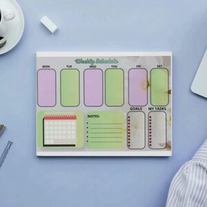 Puede incluir: Un planificador de horario semanal con un fondo gris claro. El planificador tiene secciones para cada día de la semana, notas, objetivos y tareas. Los días de la semana están etiquetados. También se ve una taza de café, un portátil y otros útiles de oficina.