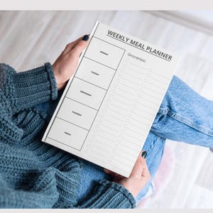 Puede incluir: Un planificador semanal de comidas blanco con el texto "WEEKLY MEAL PLANNER" en la parte superior. El planificador tiene casillas para cada día de la semana y una sección para la compra. La persona lleva un jersey verde azulado y vaqueros azules.