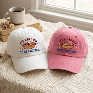 May include: Two baseball caps, one white and one pink, with the embroidered phrase "It's a bad day to be a hot dog." The caps feature a hot dog graphic with red, white, and blue stars. A coffee mug and books are in the background.