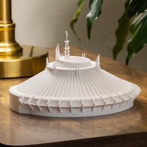 May include: A white, 3D-printed model of a futuristic structure, possibly a building or a landmark. The model has a circular base with a conical roof and detailed architectural elements. It sits on a wooden surface.