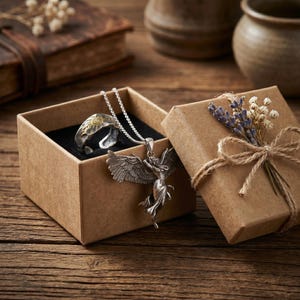 K&ouml;nnte beinhalten: Ein silberner Ring mit goldenem Sonnendesign und eine silberne Engels-Anh&auml;nger-Halskette werden in einer braunen Geschenkbox pr&auml;sentiert. Die Box ist mit Bindfaden gebunden und mit Trockenblumen verziert. Der Schmuck liegt auf einer Holzoberfl&auml;che.