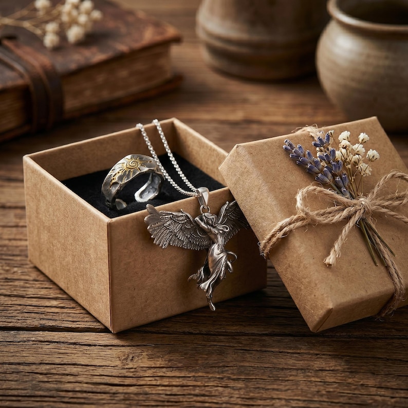 K&ouml;nnte beinhalten: Ein silberner Ring mit goldenem Sonnendesign und eine silberne Engels-Anh&auml;nger-Halskette werden in einer braunen Geschenkbox pr&auml;sentiert. Die Box ist mit Bindfaden gebunden und mit Trockenblumen verziert. Der Schmuck liegt auf einer Holzoberfl&auml;che.
