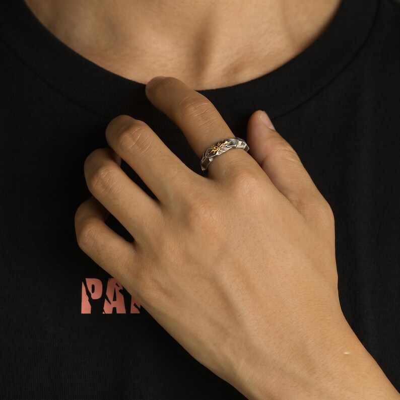 K&ouml;nnte beinhalten: Ein silberner Ring mit einem goldfarbenen floralen Design. Der Ring wird an einem Finger getragen und zeigt seine komplizierten Details und Kontrastfarben. Das Design des Rings wird vor einem dunklen Hintergrund hervorgehoben.