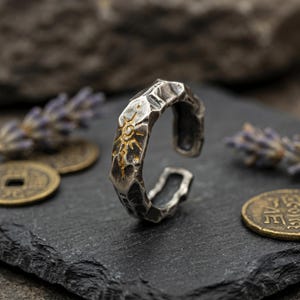 K&ouml;nnte beinhalten: Verstellbarer Ring in Silberoptik mit strukturierter Oberfl&auml;che und Sonnenmuster. Der Ring hat ein geh&auml;mmertes Aussehen und ist unten offen. Das Sonnenmuster ist goldfarben.