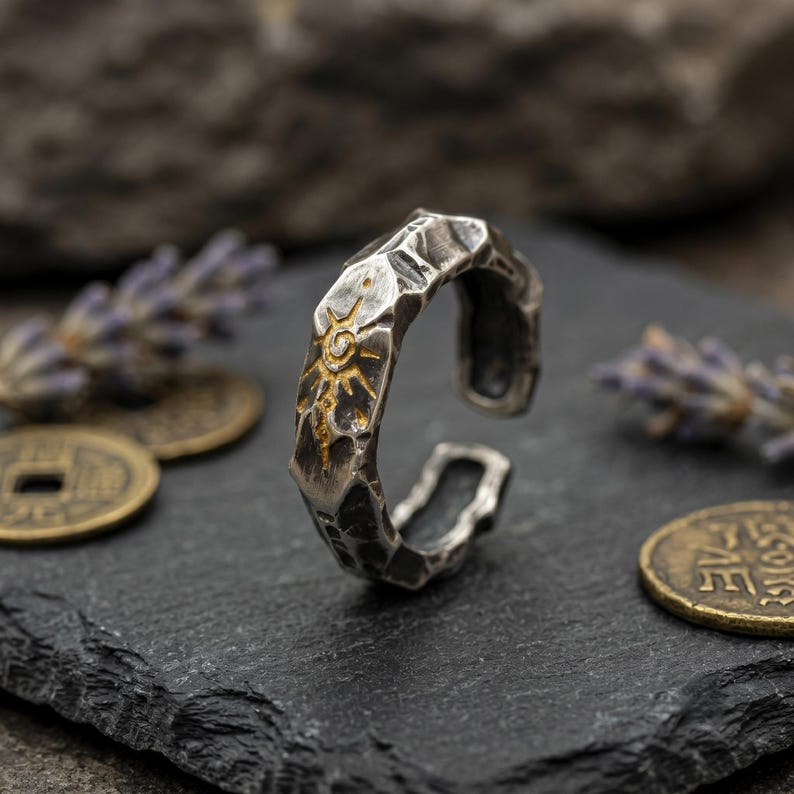 K&ouml;nnte beinhalten: Verstellbarer Ring in Silberoptik mit strukturierter Oberfl&auml;che und Sonnenmuster. Der Ring hat ein geh&auml;mmertes Aussehen und ist unten offen. Das Sonnenmuster ist goldfarben.