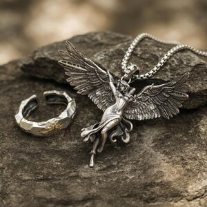 K&ouml;nnte beinhalten: Eine silberne Anh&auml;ngerkette mit einer gefl&uuml;gelten Figur mit ausgestreckten Armen und ein passender Ring mit einem Sonnenmuster. Der Schmuck wird auf einer strukturierten grauen Steinoberfl&auml;che pr&auml;sentiert, die komplizierte Details und eine Vintage-&Auml;sthetik zeigt.