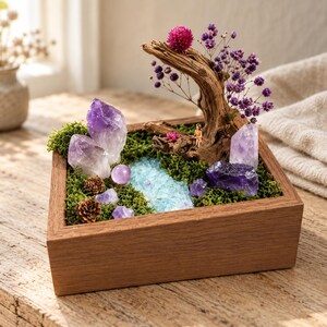 May include: A square wooden box containing a miniature landscape with green moss, purple amethyst crystal clusters, a smooth purple sphere, small pinecones, a piece of driftwood, and a small tree with purple flowers and a tiny figurine.