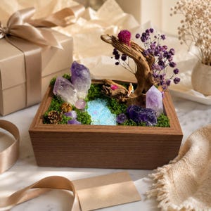 May include: A rectangular wooden box containing a miniature landscape with green moss, purple amethyst crystals, a piece of driftwood, dried purple flowers, a small blue resin stream, and a tiny figurine sitting on the wood.