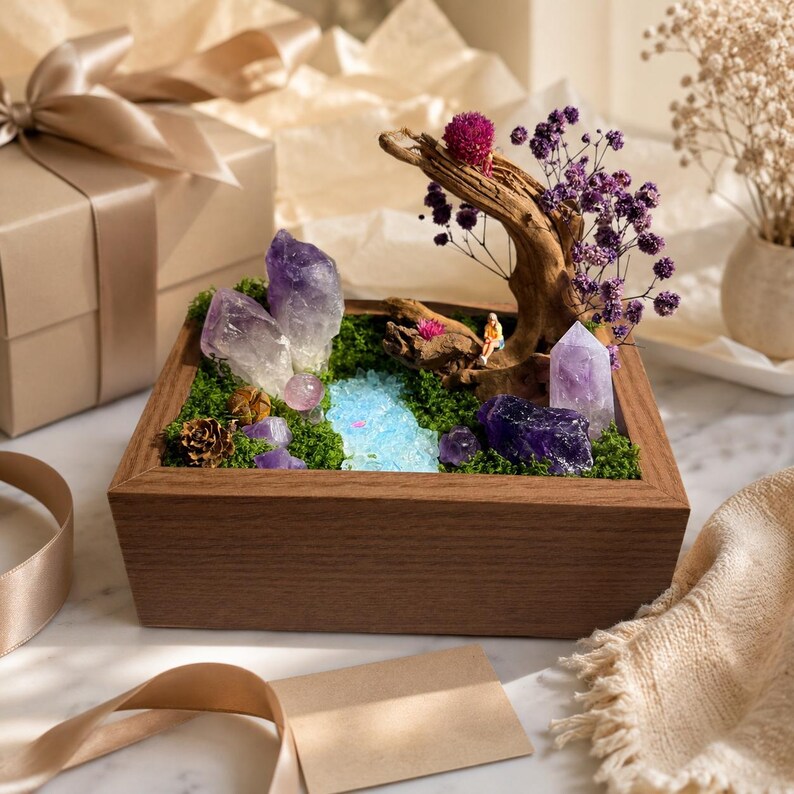 May include: A rectangular wooden box containing a miniature landscape with green moss, purple amethyst crystals, a piece of driftwood, dried purple flowers, a small blue resin stream, and a tiny figurine sitting on the wood.