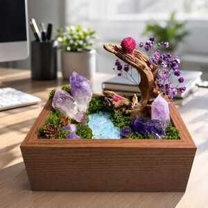 May include: A square wooden planter box containing a miniature landscape with green moss, purple amethyst crystal clusters, a piece of driftwood, dried purple flowers, a small pink flower, and a tiny figurine sitting on the wood.