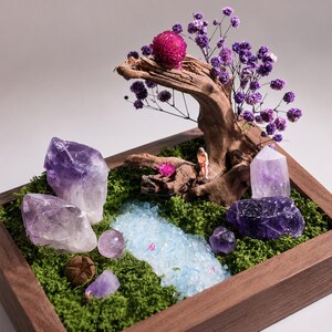 May include: A miniature landscape in a rectangular wooden frame featuring green moss, several purple amethyst crystals, a piece of driftwood, purple dried flowers, a small light blue stone path, and a tiny figurine sitting on the wood.