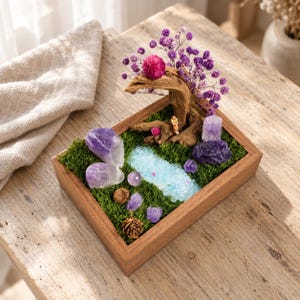 May include: A rectangular wooden tray filled with green moss, featuring a miniature landscape with a piece of driftwood, purple dried flowers, a bright pink globe amaranth, amethyst crystals, a small pinecone, and a light blue stone pond.