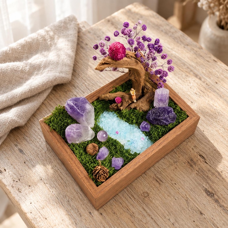 May include: A rectangular wooden tray filled with green moss, featuring a miniature landscape with a piece of driftwood, purple dried flowers, a bright pink globe amaranth, amethyst crystals, a small pinecone, and a light blue stone pond.