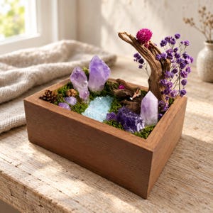May include: A rectangular wooden box containing a miniature landscape with purple amethyst crystals, green moss, a small blue stone stream, a piece of driftwood, a bright pink globe amaranth flower, and sprigs of purple statice flowers.