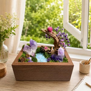 May include: A square wooden planter box containing a miniature landscape with green moss, light blue resin water, purple amethyst crystal clusters, a piece of driftwood, and small purple dried flowers. A tiny figurine sits on the wood.