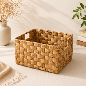 Puede incluir: Una cesta de almacenamiento rectangular hecha de fibras naturales tejidas en un tono marrón claro. La cesta presenta un patrón de tejido de damero consistente en sus lados y base, con asas recortadas integradas en dos lados opuestos.