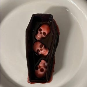 May include: A dark brown, coffin-shaped container filled with a dark liquid and three small, pale pink, skull-shaped objects submerged inside. The coffin has a smooth, molded exterior and is positioned centrally on a plain white surface.