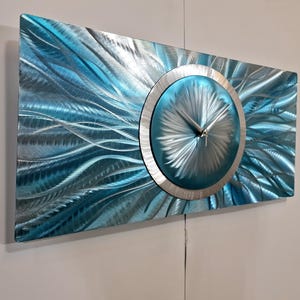 Puede incluir: Un reloj de pared moderno con una esfera rectangular de metal cepillado en tonos azules y plateados. El reloj presenta una esfera circular con manecillas plateadas y un marco de metal cepillado. El diseño tiene un patrón texturizado y fluido.