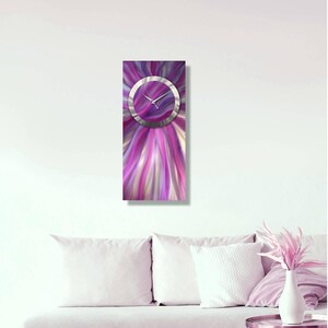 Puede incluir: Un reloj de pared rectangular con un diseño abstracto vibrante en tonos de morado, rosa y blanco. La esfera del reloj es un círculo plateado con manecillas plateadas. El reloj está montado en una pared blanca.