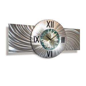 Puede incluir: Un reloj de pared moderno de metal plateado con números romanos. La esfera del reloj presenta un centro azul claro con un diseño texturizado. El reloj es rectangular con una esfera circular en el centro.