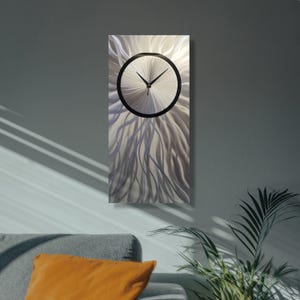 Puede incluir: Un reloj de pared rectangular de metal con un diseño plateado y negro. La esfera del reloj es un círculo con manecillas negras y un borde negro. El fondo tiene un patrón texturizado y fluido.
