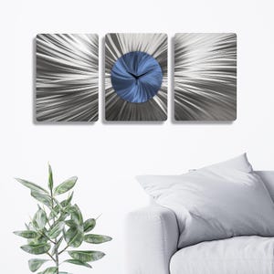 Puede incluir: Un reloj de pared de tres paneles con un diseño moderno. El reloj presenta un centro circular azul con manecillas negras, sobre un patrón radial plateado y gris. Los paneles rectangulares están montados en una pared blanca.