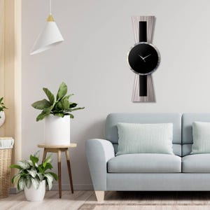 Puede incluir: Un reloj de pared moderno con esfera negra y manecillas plateadas. El reloj está colocado sobre un diseño vertical con detalles plateados y negros. El reloj está montado en una pared blanca sobre un sofá azul claro. La habitación está decorada con plantas.