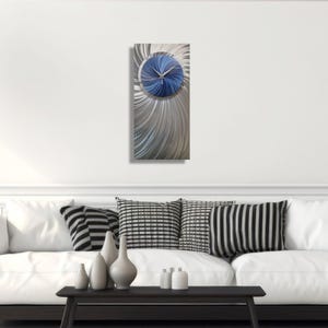 Puede incluir: Un reloj de pared moderno con una esfera circular azul y un diseño radial plateado. Las manecillas del reloj son negras. El reloj rectangular mide aproximadamente 76 cm de alto y está colgado en una pared blanca.