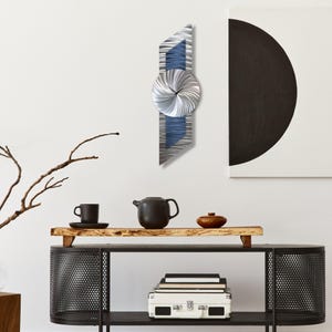 Puede incluir: Reloj de pared moderno de metal con diseño geométrico plateado, azul y gris. El reloj tiene una esfera circular con un patrón en espiral y un cuerpo rectangular. Está montado en una pared blanca, encima de una estantería negra.