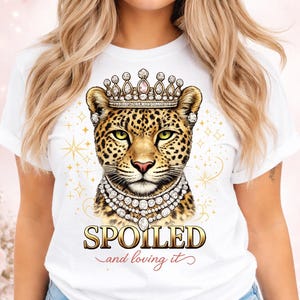 May include: White t-shirt featuring a leopard wearing a crown and a diamond necklace. The leopard's face is detailed with spots and green eyes. The text "SPOILED and loving it" is printed below the leopard.