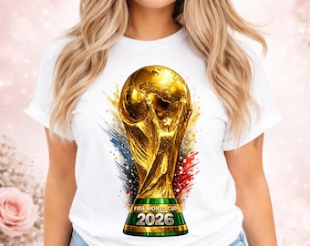 Op de afbeelding: Wit T-shirt met een gouden FIFA World Cup trofee ontwerp. De trofee staat tegen een achtergrond van blauwe, rode en gele penseelstreken. De tekst "FIFA WORLD CUP 2026" staat op de basis van de trofee.