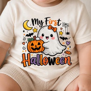 May include: A cream-colored short-sleeve t-shirt featuring a graphic of a white ghost with an orange bow holding an orange jack-o'-lantern. The design includes a yellow crescent moon, stars, bats, a spider web, and the text My First Halloween.