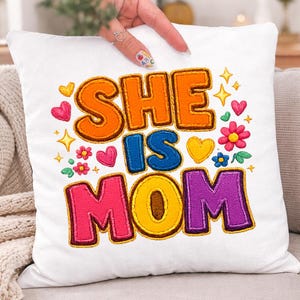 Könnte beinhalten: Weißes quadratisches Kissen mit dem gestickten Text "SHE IS MOM" in Orange, Blau, Rosa und Lila. Das Design umfasst Herzen, Blumen und Sterne in verschiedenen Farben. Das Kissen liegt auf einem grauen Sofa.