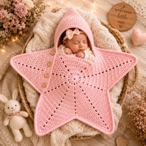 Könnte beinhalten: Eine hellrosa gehäkelte sternförmige Baby-Wickeldecke mit spitzer Kapuze und vier Holzknöpfen an einer Seite. Die Wickeldecke weist ein strukturiertes Strickmuster auf und ist so konzipiert, dass sie ein Kleinkind warm und sicher umschließt.