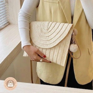 Könnte beinhalten: Beige Stroh-Clutch mit Klappenverschluss und kleinem Bommeldetail. Die Clutch hat eine gewebte Textur und einen langen, dünnen Riemen. Die Person trägt ein weißes Langarmshirt und eine gelbe Weste.