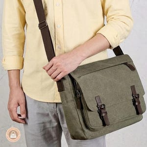 May include: An olive green canvas messenger bag with a dark brown adjustable shoulder strap. The bag features a front flap secured by two dark brown leather-like buckle straps, metal hardware, and a zippered compartment under the main flap.
