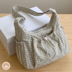 May include: A beige and white gingham patterned shoulder bag featuring a main zippered compartment, a front pocket with a drawstring toggle closure, and an adjustable shoulder strap. The bag has a soft, unstructured design with side pockets.