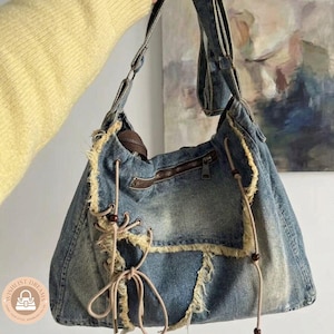 May include: A denim shoulder bag with a distressed, faded blue wash. The bag features a brown zipper, frayed edges, and decorative beige ties with wooden beads. The bag has a long strap and a large capacity.