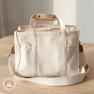 Puede incluir: Un bolso tote de lona color crema con forma rectangular estructurada, dos asas superiores con agarre de cuero color canela y una correa de hombro ajustable. El exterior incluye múltiples bolsillos abiertos y costuras verticales reforzadas.
