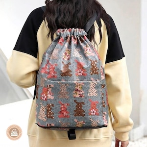 May include: A grey drawstring backpack featuring a repeating pattern of stylised rabbits in various shades of pink, brown, and cream with floral and polka dot details. The bag has a gathered top closure with black straps and a horizontal front pocket.