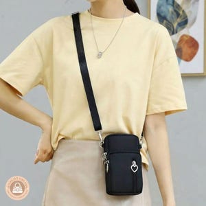 May include: A black rectangular crossbody bag with a smooth finish, featuring a vertical zippered front pocket with a silver heart-shaped zipper pull. The bag includes a wide black adjustable strap and is worn over a pale yellow t-shirt and skirt.