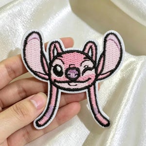 以下が含まれることがあります： 大きな耳、ウィンクした目、小さな鼻を持つ、ピンク色のキャラクターが描かれた刺繍ワッペン。デザインは黒い糸で縁取られており、耳と顔の部分は淡いピンク色で埋め尽くされており、アニメのような独特の見た目をしています。
