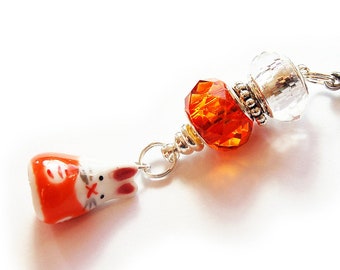 May include: A charm featuring a white and orange ceramic bunny with red accents, connected to a chain with orange and clear faceted beads. The charm has a silver-coloured clasp.