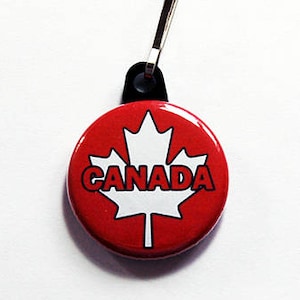 Könnte beinhalten: Ein runder, roter Knopf mit einem weißen Ahornblatt und dem Wort "CANADA" in Weiß. Der Knopf ist an einer schwarzen Schlaufe und einem silberfarbenen Metallclip befestigt.
