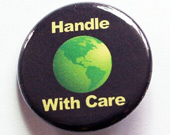 Könnte beinhalten: Ein runder, schwarzer Button mit den gelben Worten "Handle With Care". Der Button zeigt eine grüne Weltkugel-Grafik in der Mitte. Der Button ist vor einem weißen Hintergrund platziert.