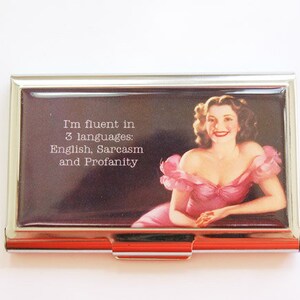 Può includere: Un porta carte di credito argentato con un'immagine vintage di una donna in abito rosa. Il testo sul porta carte recita: "I'm fluent in 3 languages: English, Sarcasm and Profanity."
