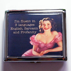 Può includere: Un compatto in metallo argentato con sfondo blu scuro con un'illustrazione vintage di una donna in abito rosa. Il testo recita: "Parlo fluentemente 3 lingue: Inglese, Sarcasmo e Profanità."