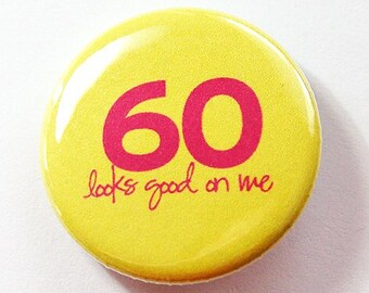 May include: A bright yellow button badge with the number "60" in pink, and the words "looks good on me" in pink script. The badge is round and shiny. A celebratory accessory for a 60th birthday.