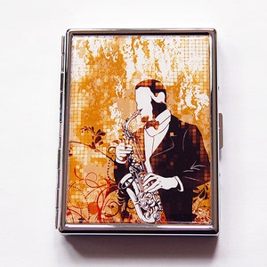 May include: A silver cigarette case with a design of a jazz musician playing a saxophone. The background is brown and orange with floral and geometric patterns. The musician is depicted in a tuxedo and bow tie.