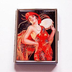 Puede incluir: Un estuche de cigarrillos de metal plateado con una ilustración vintage de una mujer con un vestido y sombrero rojos, sosteniendo una pandereta. La obra de arte tiene un fondo negro con confeti y una máscara roja.